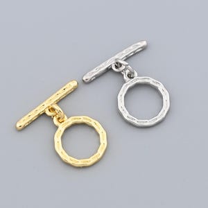 May include: Two toggle clasps, one gold-coloured and one silver-coloured, for jewellery making. Each clasp features a textured bar and a textured circular ring. The clasps are displayed against a solid grey background.