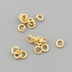 May include: A collection of small, gold-colored jewelry clasps and connectors. The clasps are in various shapes, including circular and interlocking designs. The pieces are scattered on a gray surface, showcasing their intricate details and shiny finish.