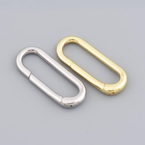 Può includere: Due chiusure metalliche di forma ovale, una argentata e una dorata, sono esposte su uno sfondo grigio. Le chiusure hanno una forma simile, con un design rettangolare arrotondato. La chiusura argentata è a sinistra e quella dorata a destra.