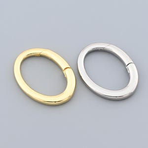 May include: Two oval metal clasps, one gold and one silver, are shown against a grey background. The clasps are closed, with a small gap where they connect. The design is simple and modern, suitable for jewellery or accessories.