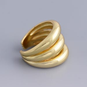 Puede incluir: Un anillo dorado con un diseño en capas. El anillo presenta tres bandas curvas que crean un efecto apilado. El anillo está hecho de un metal brillante y reflectante, sobre un fondo gris.