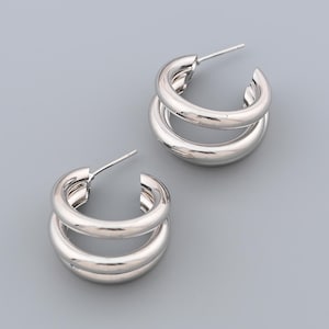 May include: A pair of silver-colored double hoop earrings. Each earring features two connected, rounded hoops. The earrings are displayed against a solid gray background, highlighting their polished, metallic finish. The design is simple and modern.