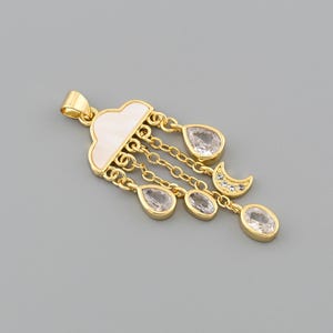 Op de afbeelding: Een goudkleurige hanger met een wolkenontwerp, witte inleg en hangende, druppelvormige, heldere stenen. Een maansikkelsieraad en extra heldere stenen zijn ook inbegrepen. De hanger ligt op een grijze achtergrond.