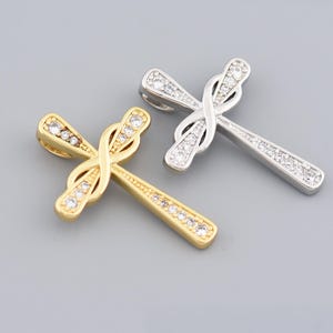 Puede incluir: Dos colgantes de cruz, uno dorado y otro plateado, cada uno adornado con brillantes gemas transparentes. Las cruces presentan un diseño de símbolo de infinito en el centro, sobre un fondo gris.