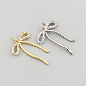 May include: Two bow-shaped pendants. One is gold-toned with clear stones on the bow, the other is silver-toned with clear stones on the bow. Both have curved, dangling elements.