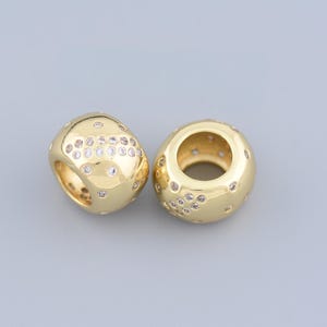 May include: Two gold-colored, rounded beads with a hollow center. Each bead is embellished with small, clear, round gemstones. The beads have a smooth, polished finish and are displayed against a gray background.