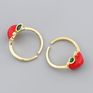 May include: Two gold-colored adjustable rings, each adorned with a red enamel apple and a green enamel leaf. The rings are set against a gray background, showcasing the jewelry's design.