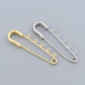 May include: Two large safety pins, one gold-colored and one silver-colored, are displayed against a gray background. Each pin has a series of small loops along the pin's shaft, designed for attaching charms or other decorative elements.