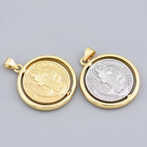 Puede incluir: Dos colgantes de San Benito en tono dorado y plateado. Cada colgante presenta un diseño circular con un emblema religioso detallado. Los colgantes tienen una anilla dorada para sujetar a una cadena. El fondo es gris claro liso.