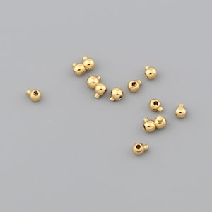 Peut inclure: Plusieurs petites perles dorées et brillantes sont dispersées sur une surface grise. Chaque perle est ronde avec un petit trou, suggérant qu'elles sont destinées à la fabrication de bijoux ou à l'artisanat. Les perles varient légèrement en taille.