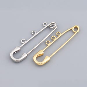 May include: Two metal safety pins, one silver and one gold, are displayed against a gray background. Each pin has a classic safety pin design with a curved end and a clasp. Both pins have three small loops for attaching charms or other embellishments.