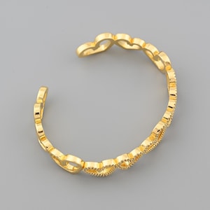 Könnte beinhalten: Ein goldfarbenes Armreif-Armband mit einem sich wiederholenden herzförmigen Design. Das offene Armband hat eine polierte Oberfläche und ist vor einem grauen Hintergrund platziert. Das Design des Armbands ist offen und luftig, wobei jedes Herz mit dem nächsten verbunden ist.