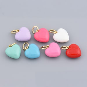 May include: A collection of eight heart-shaped charms in various colors, including lavender, red, white, pink, blue, and teal. Each charm has a gold-colored loop for attaching to jewelry or other crafts. The charms are arranged on a gray surface.