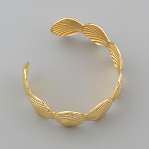 May include: A gold-colored cuff bracelet with a seashell design. The bracelet has an open design and is made of a shiny, metallic material. The seashell design is repeated around the bracelet.
