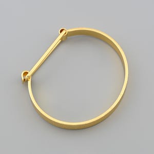 May include: A gold-colored bangle bracelet with a straight bar closure. The bracelet has a textured surface and a simple, elegant design. The jewelry is shown against a gray background.