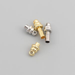 Op de afbeelding: Een verzameling van vier kleine metalen koordeinden voor sieraden, bestaande uit twee goudkleurige en twee zilverkleurige onderdelen. Elk onderdeel heeft een afgerond, getrapt bovendeel met een klein oogje en een holle cilindrische basis.