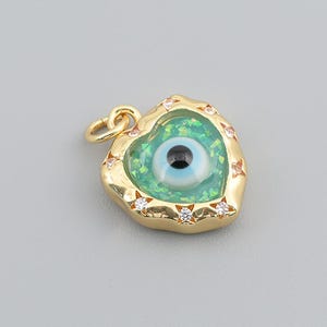 May include: A gold-toned heart-shaped pendant featuring a central evil eye motif set against a shimmering green background. The metallic frame is accented with small, clear, round-cut stones embedded along the edge and includes a small jump ring.