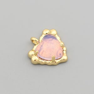 May include: A small, heart-shaped pendant with a light pink gemstone center. The gemstone is encased in a gold-colored metal frame with a textured, irregular border. A small loop is attached for hanging. The pendant is set against a neutral gray background.