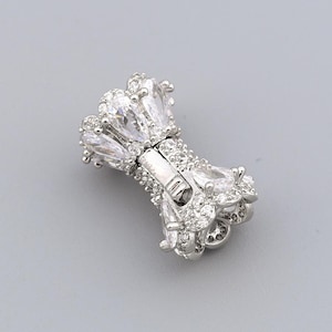 May include: A silver-toned bow-shaped jewelry clasp with clear, sparkling crystals. The clasp features a central bar with a hinged closure and is adorned with multiple crystal accents, creating a glamorous appearance. The background is a solid, light gray.