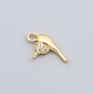 Puede incluir: Un colgante dorado con forma de pájaro estilizado, con un lazo para una cadena. El cuerpo del pájaro está adornado con pequeños cristales transparentes, que añaden brillo. El colgante está sobre un fondo gris liso.