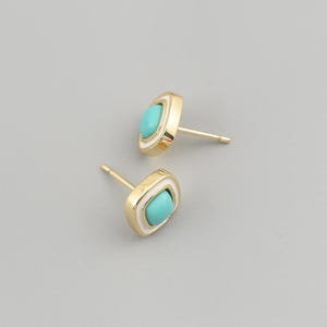 May include: A pair of gold stud earrings featuring a square-shaped frame with rounded corners. Each earring contains a central, smooth, bright turquoise stone surrounded by a thin white border, set within a polished gold-tone metal base.