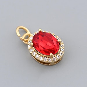 Puede incluir: Colgante dorado con una gema roja brillante de forma ovalada en el centro. La gema está rodeada por un halo de pequeños cristales transparentes. El colgante tiene un pequeño lazo en la parte superior.