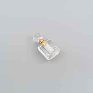 May include: A small, clear glass perfume bottle with a rectangular base and a faceted, rounded stopper. The neck of the bottle features a gold-colored metal band and a white cylindrical insert, creating a simple and functional design.