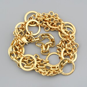 May include: A gold-colored necklace with a variety of chain links and large circular rings. The necklace is arranged in a circular pattern against a gray background. The chain links are of varying sizes and shapes.