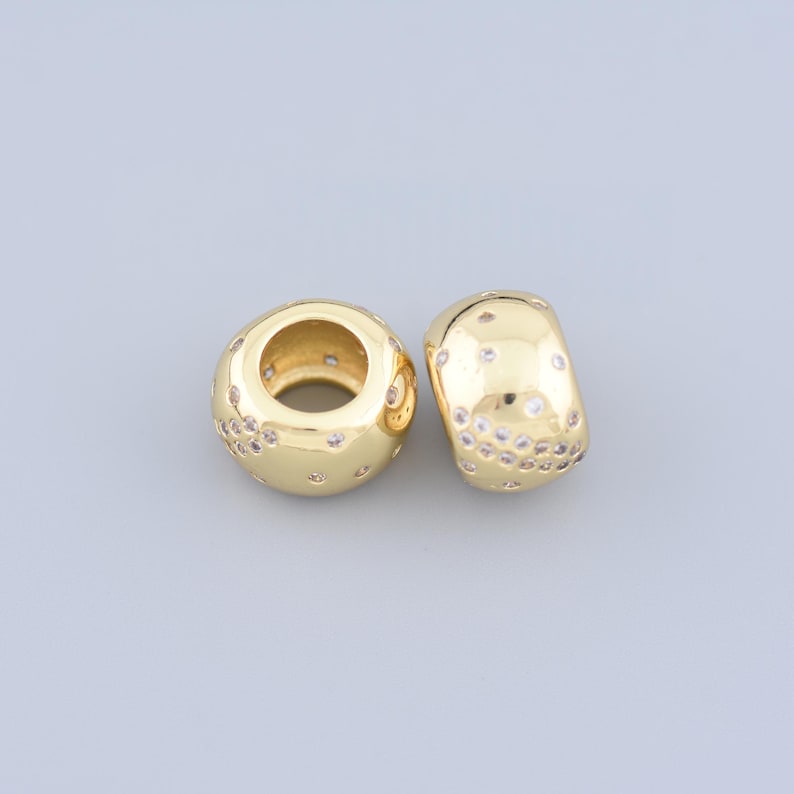 May include: Two gold-colored beads with a polished finish. Each bead is adorned with small, clear, sparkling accents. One bead is round, and the other is a barrel shape. These beads are suitable for jewelry making.