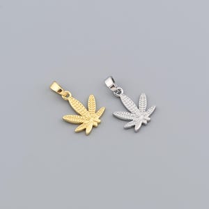 May include: Two pendant necklaces, one gold-colored and one silver-colored, each shaped like a cannabis leaf. The pendants have a bail at the top for a chain. The background is a solid, light gray color.