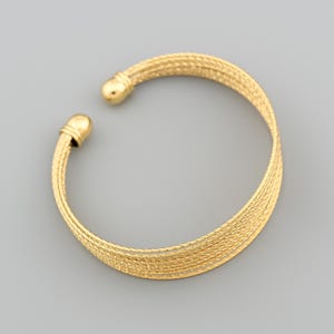 Könnte beinhalten: Ein goldfarbenes Armreif-Armband mit einem strukturierten, gewebten Design. Das Armband hat eine abgerundete Form mit einem kleinen, kugelförmigen Detail an jedem Ende. Das offene Design ermöglicht ein einfaches An- und Ausziehen. Das Armband besteht aus glänzendem Metall.
