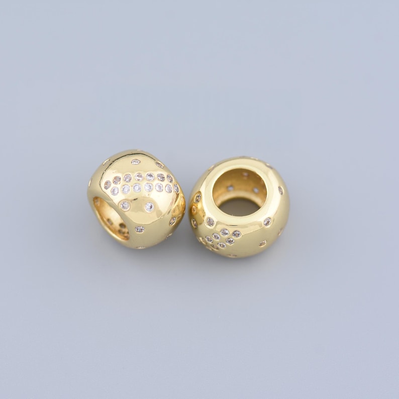 May include: Two gold-colored, rounded beads with a hollow center. Each bead is embellished with small, clear, round gemstones. The beads have a smooth, polished finish and are displayed against a gray background.