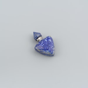 Puede incluir: Un pequeño frasco de perfume triangular tallado en piedra lapislázuli de color azul intenso con motas blancas. El frasco presenta una superficie facetada y un tapón de metal plateado puntiagudo en la parte superior, unido al cuerpo.