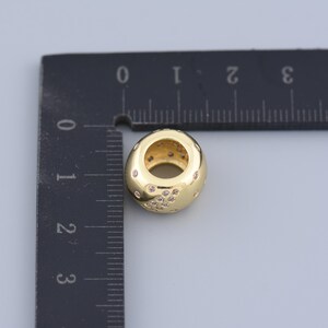 May include: A gold-colored bead with a central hole and small, clear embellishments. The bead is oval-shaped and sits next to a ruler, which measures the bead at approximately 0.5 inches in length.