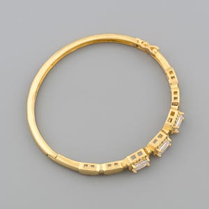 May include: A gold-colored bangle bracelet with a series of rectangular, clear gemstones. The bracelet has a hinged clasp and a simple, elegant design. The gemstones are set in a row along the front of the bracelet.