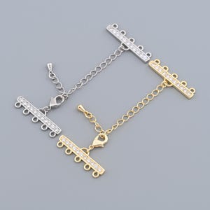 Puede incluir: Dos cierres de collar de múltiples hilos, uno plateado y otro dorado, cada uno con una barra de pedrería transparente. Los cierres tienen múltiples bucles para sujetar los hilos y una extensión de cadena.