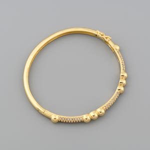 May include: A gold-colored bangle bracelet with a hinged clasp. The bracelet features a row of small, clear stones and several rounded gold beads. The design is simple and elegant, suitable for everyday wear or special occasions.