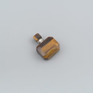 May include: A small, rectangular pendant crafted from polished tiger's eye gemstone, featuring a distinct golden-brown striped pattern. The stone is faceted with beveled edges and is attached to a small, silver-toned metal bail at the top.
