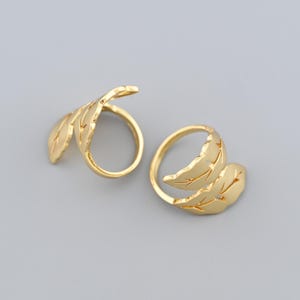 May include: Two gold-colored leaf-shaped rings. Each ring features a delicate leaf design that wraps around the finger. The rings are set against a gray background, highlighting their intricate details and shiny finish.