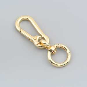 May include: A gold-toned metal lobster claw clasp featuring an elongated, teardrop-shaped hook connected to a circular swivel ring. The hardware has a polished, reflective finish and a spring-loaded gate mechanism on the main hook.