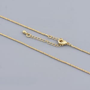 May include: A delicate gold necklace with a textured chain and a lobster clasp closure. The necklace has an adjustable chain for a customized fit. The jewelry is displayed against a solid grey background.