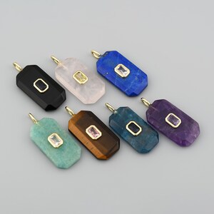 May include: A collection of gemstone pendants in various colors, including black, pink, blue, and purple. Each pendant has a rectangular shape with a gold-colored bail and a smaller, contrasting gemstone set in the center.
