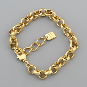 May include: A gold-colored bracelet with a chunky chain link design. The bracelet features a lobster clasp closure and a small rectangular charm with the words "Good Luck" engraved on it. The bracelet is displayed on a gray background.