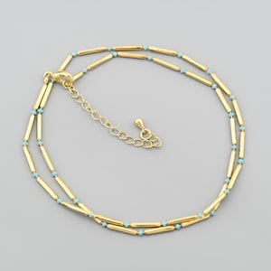 May include: Gold-colored anklet with a double-strand design. The anklet features small, light blue beads interspersed between gold-colored, elongated beads. The clasp and adjustable chain are also gold-colored, creating a cohesive and elegant look.