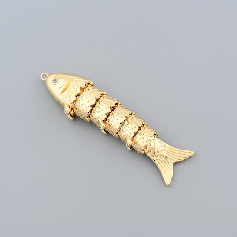 May include: A gold-colored articulated fish pendant with a small, clear gem on its head. The fish has detailed scales and fins, and a loop for attaching to a chain or other jewelry. The pendant is made of metal.