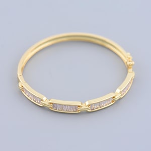 May include: A gold-colored bangle bracelet with rectangular, clear gemstones set in a repeating pattern. The bracelet has a hinged clasp and a smooth, polished finish. The background is a solid, light gray.