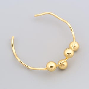 Op de afbeelding: Een goudkleurige manchetarmband met een golvend ontwerp. De armband heeft drie bolvormige gouden kralen. Het open ontwerp maakt het gemakkelijk om te dragen. De sieraden zijn geplaatst tegen een lichtgrijze achtergrond.