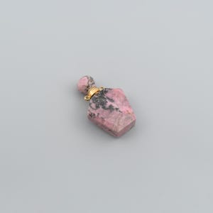 May include: A small perfume bottle pendant crafted from pink and black rhodonite stone. The bottle features a faceted, shield-shaped body with a matching round stone stopper and a gold-toned metal collar with two small loops for attachment.
