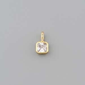 Peut inclure: Un pendentif dor&eacute; avec une pierre pr&eacute;cieuse carr&eacute;e et transparente. Le pendentif a une petite boucle en haut, incrust&eacute;e de minuscules pierres scintillantes. La pierre est sertie dans une lunette dor&eacute;e.