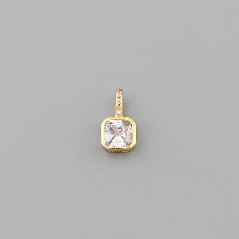 Peut inclure: Un pendentif dor&eacute; avec une pierre pr&eacute;cieuse carr&eacute;e et transparente. Le pendentif a une petite boucle en haut, incrust&eacute;e de minuscules pierres scintillantes. La pierre est sertie dans une lunette dor&eacute;e.